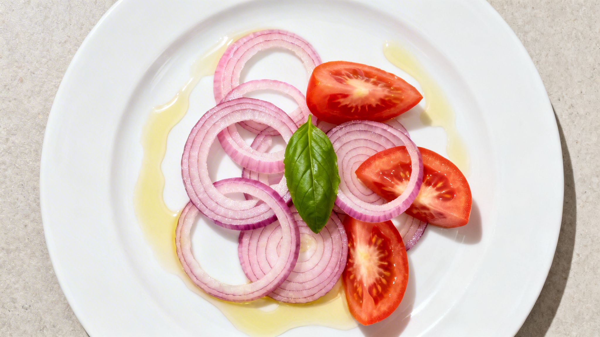 Plato blanco con aros de cebolla morada, gajos de tomate, hoja de albahaca y aceite de oliva.