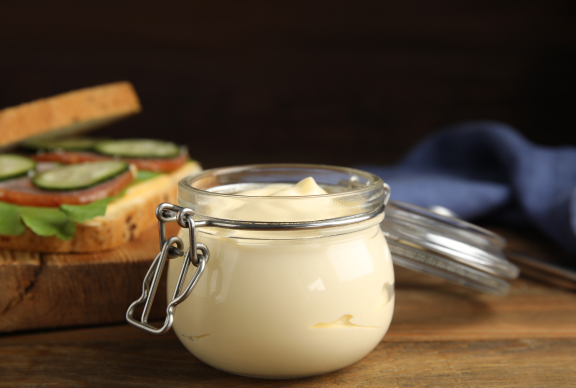 Tarro de mayonesa casera de textura cremosa, preparada con yemas de huevo pasteurizadas, sobre una mesa de madera junto a una tostada con vegetales.
